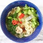 Photo of a blue bowl with noodle soup and vegetables against a marble background