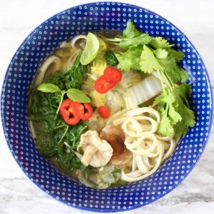 Photo of a blue bowl with noodle soup and vegetables against a marble background