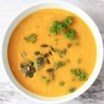 Photo of smooth orange pumpkin soup topped with green pumpkin seeds and curly parsley in a light blue bowl against a marble background