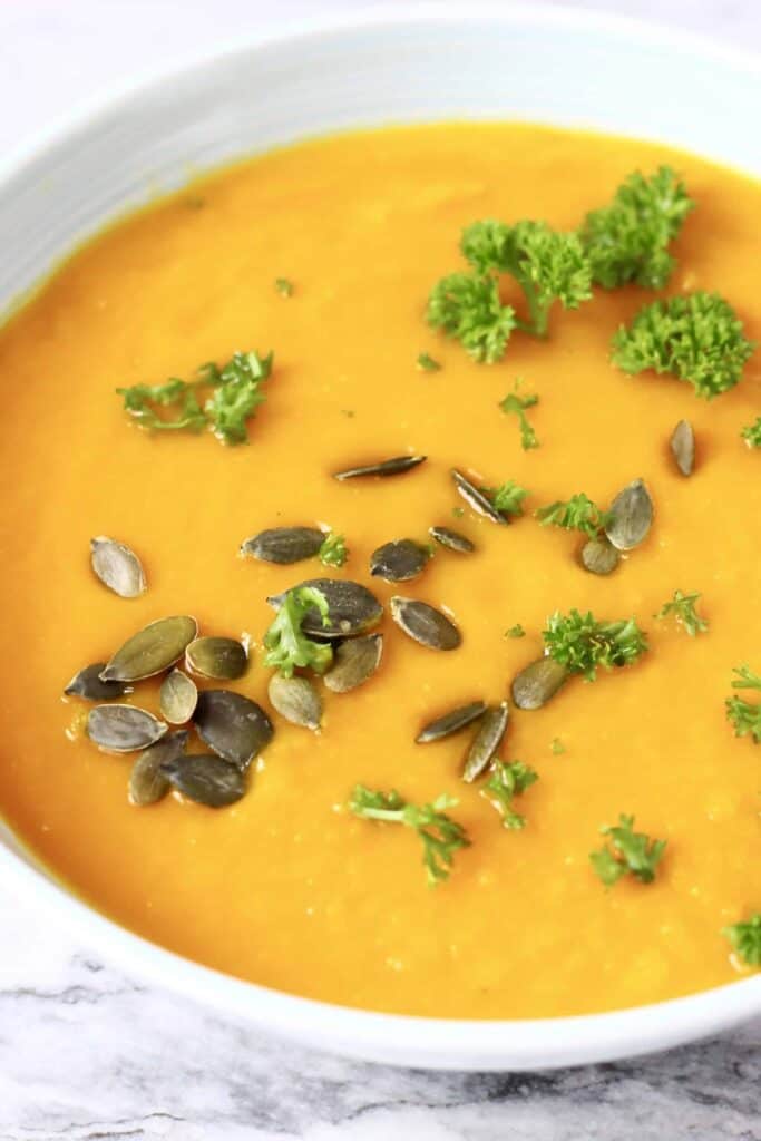 Photo of smooth orange pumpkin soup topped with green pumpkin seeds and curly parsley in a light blue bowl against a marble background