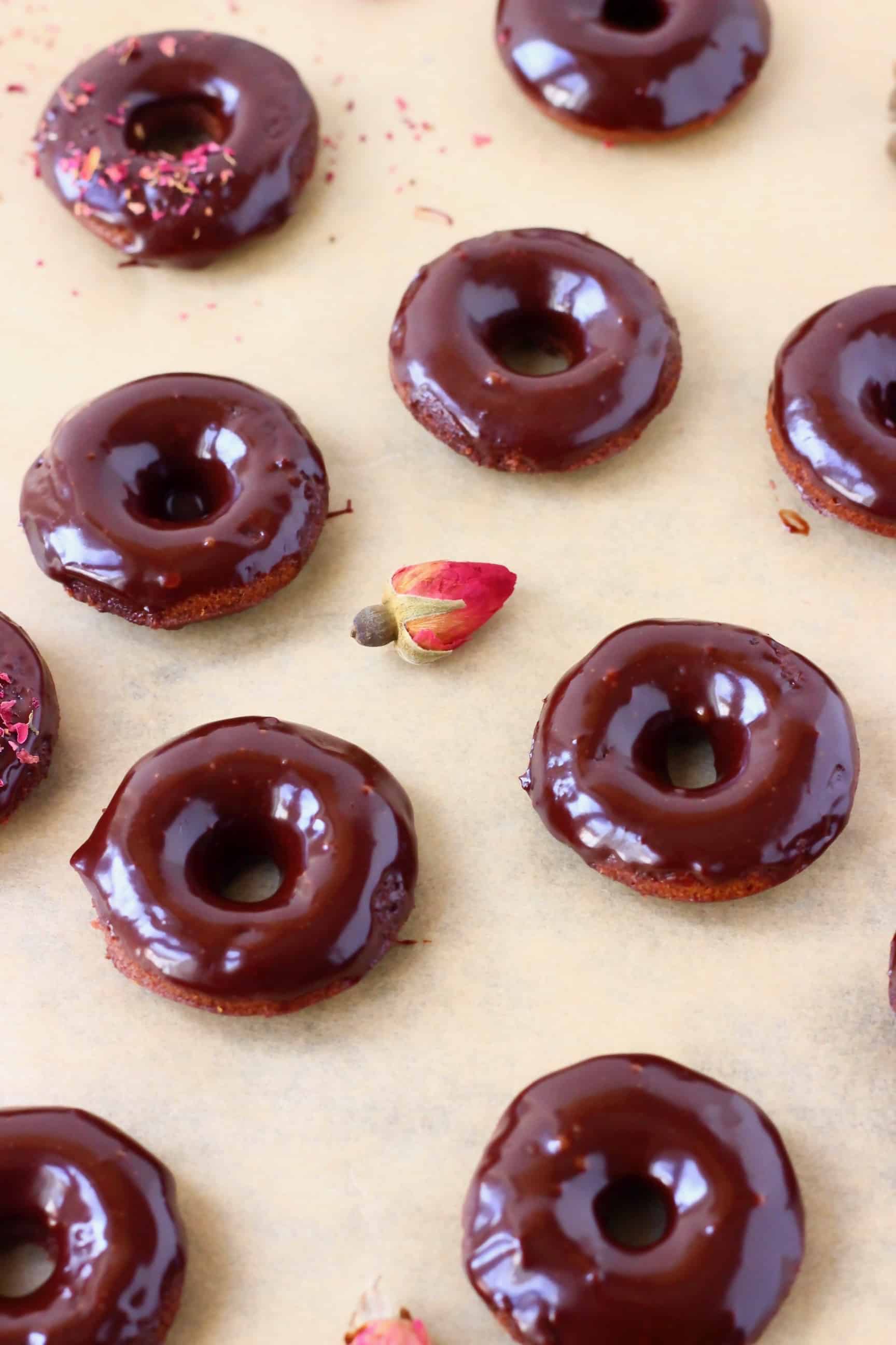 These Vegan Chocolate Baked Donuts are moist and fluffy and covered in a shiny chocolate glaze. A perfect snack or dessert for parties! Egg-free, dairy-free, gluten-free and refined sugar free. #rhiansrecipes #donuts #vegan #glutenfree #dairyfree Chocolate glazed mini chocolate donuts on a sheet of brown baking paper decorated with dried roses