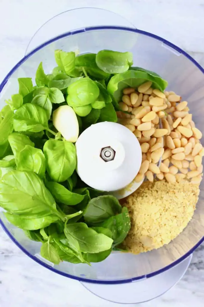 Fresh basil, garlic, nutritional yeast and pine nuts in a food processor against a marble background