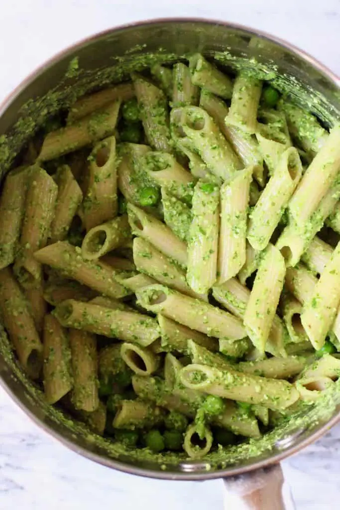 Cooked penne pasta and green peas in a silver saucepan with green pesto