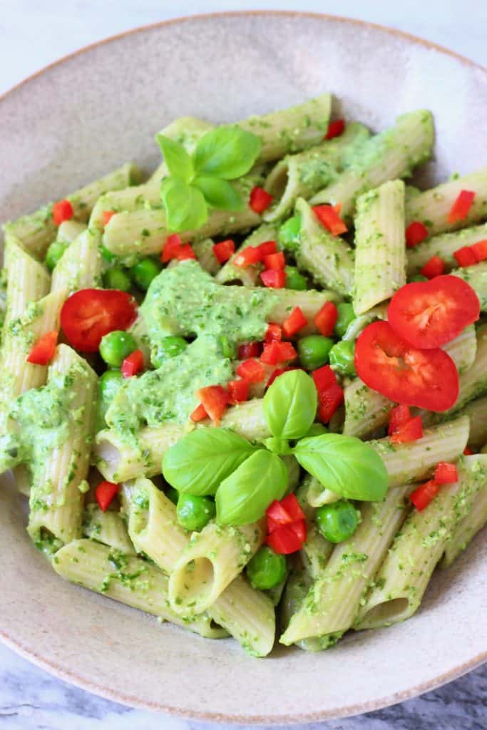 This Vegan Pesto Pasta is super easy to make, nutty and fragrant, and easily customisable! Includes a recipe for homemade vegan pesto. An easy plant-based dinner. Vegetarian, dairy-free, egg-free, gluten-free. #rhiansrecipes #pesto #pasta #vegan #glutenfree #dairyfree Penne pasta with pesto sauce, green peas and chopped red pepper in a beige bowl against a marble background