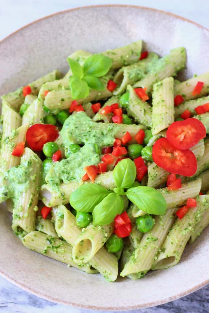 Penne pasta with pesto sauce, green peas and chopped red pepper in a beige bowl against a marble background