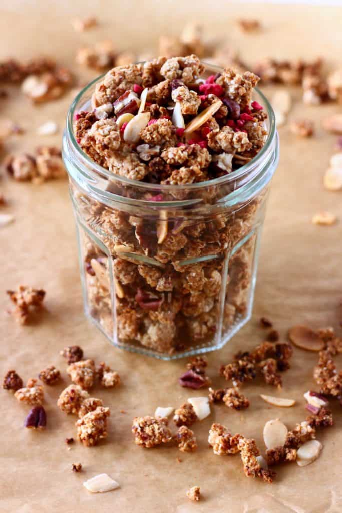 Granola with pecan nuts and flaked almonds in a glass jar on a sheet of brown baking paper scattered with granola pieces