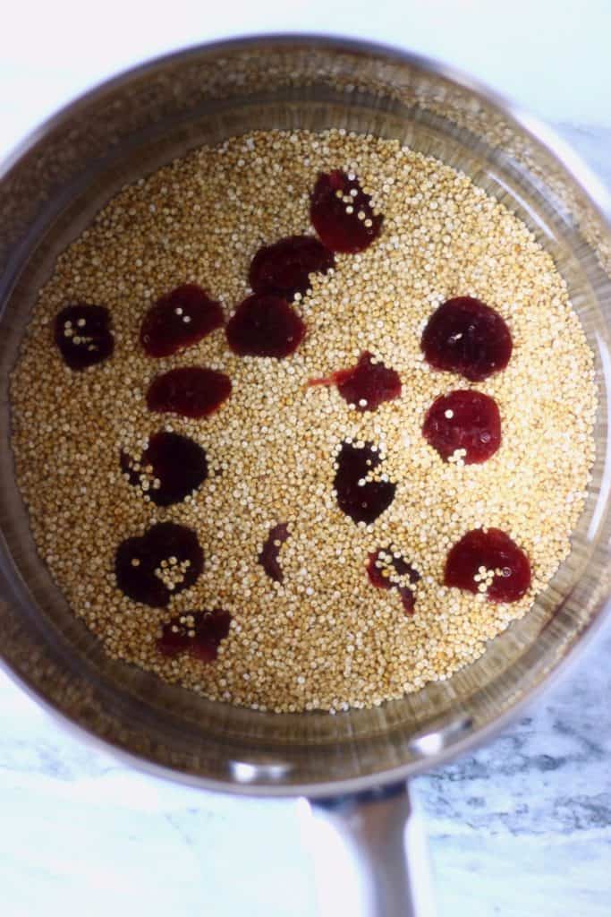 Raw quinoa, dried cranberries and water in a silver saucepan against a marble background