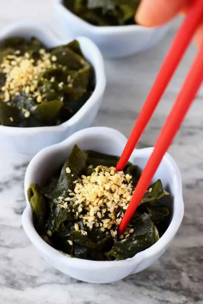 Seaweed salad topped with sesame seeds in three small blue bowls against a marble background with a pair of red chopsticks stuck in one of the bowls