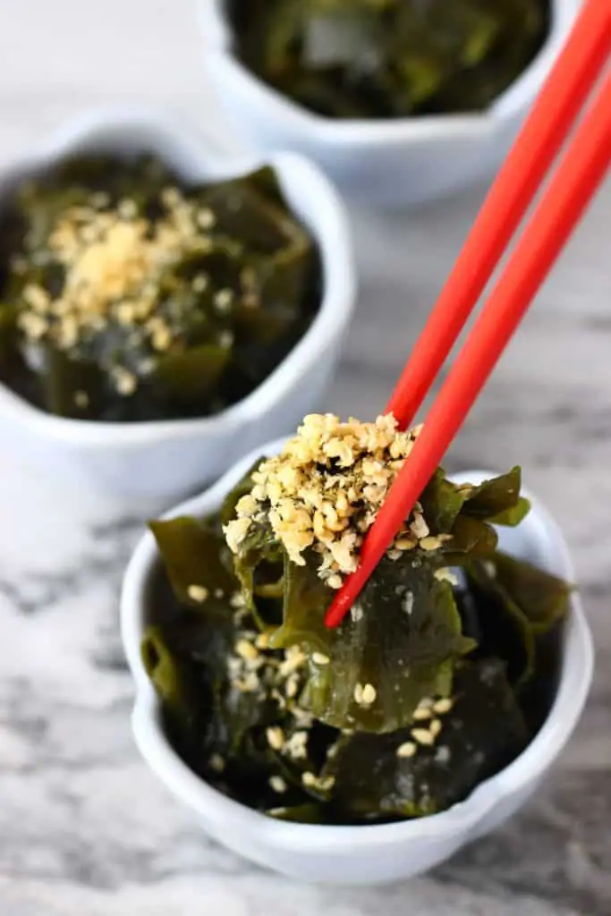 Seaweed salad topped with sesame seeds in three small blue bowls against a marble background with a pair of red chopsticks stuck in one of the bowls