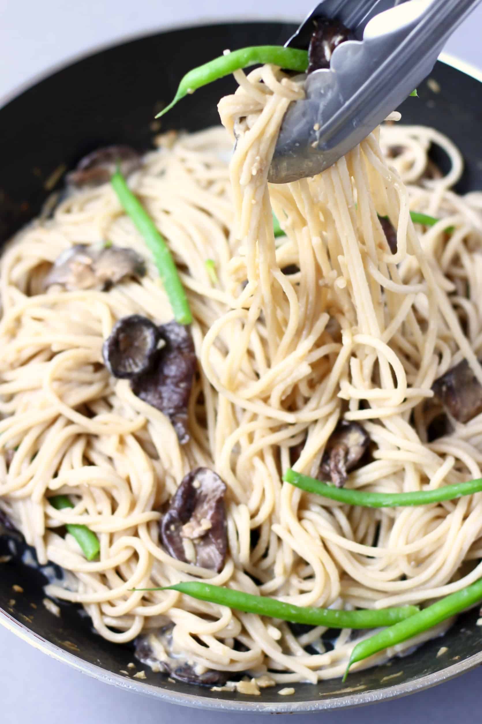 Spaghetti with creamy vegan miso pasta sauce, shiitake mushrooms and French beans being lifted up with tongs in a black frying pan 