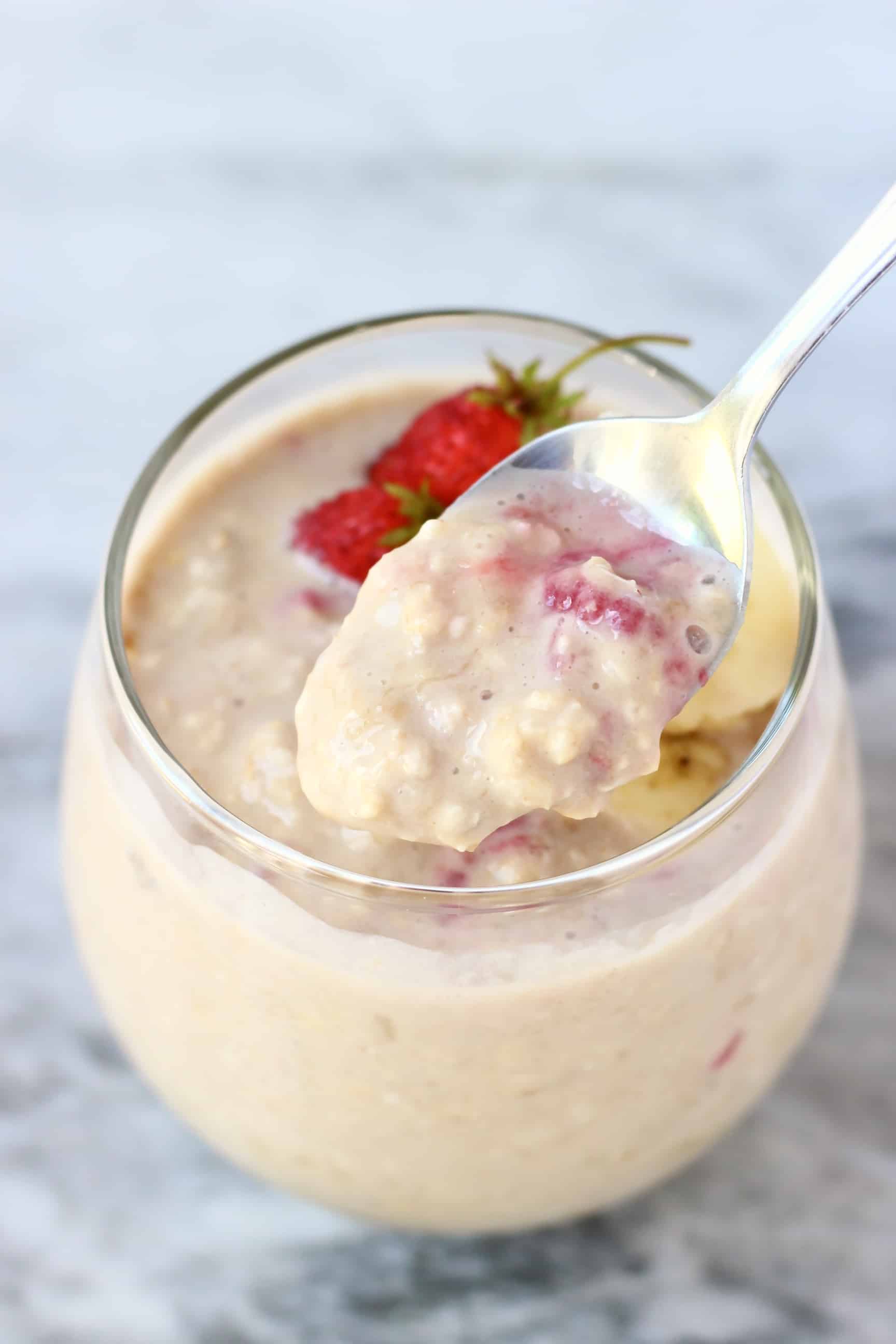 Banana overnight oats in a glass cup topped with sliced bananas and mini strawberries with a spoon holding up a mouthful