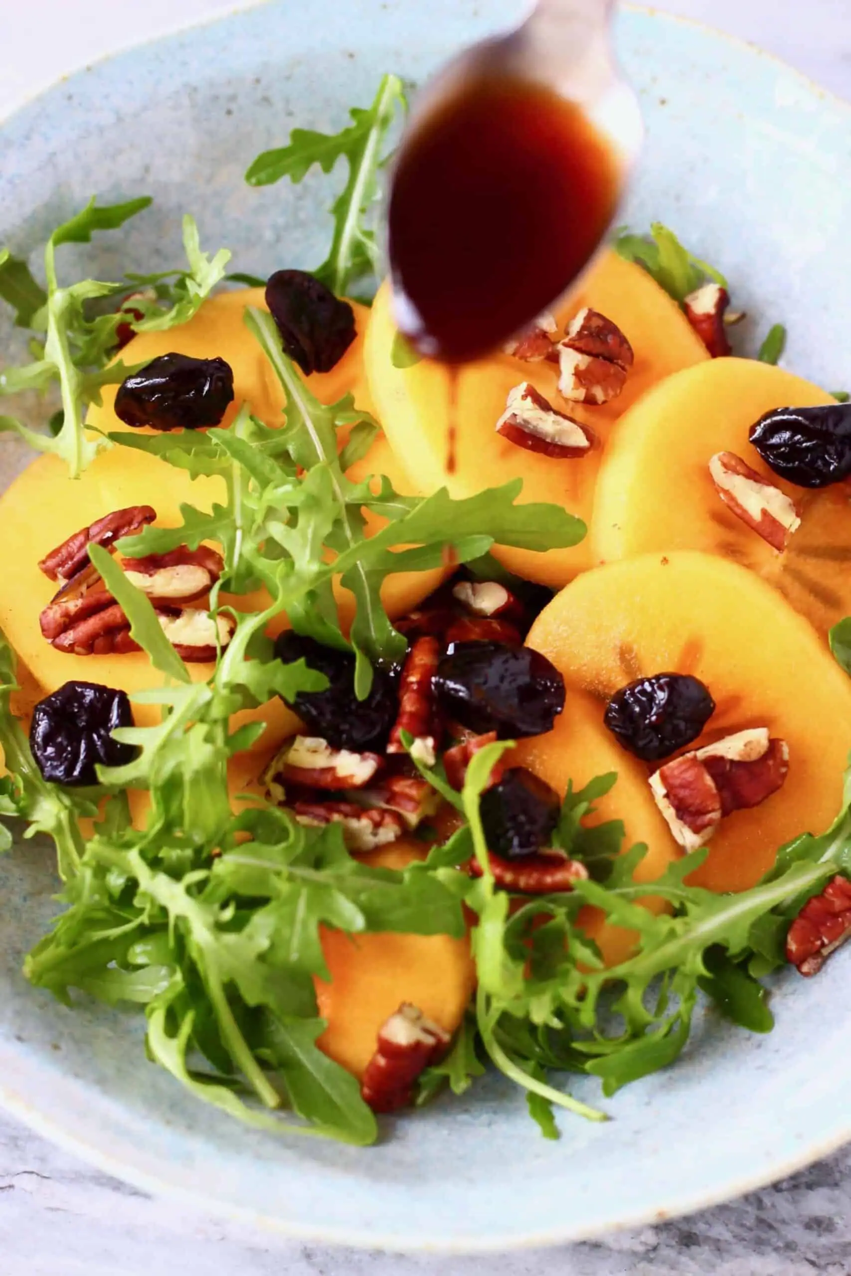 Persimmon salad with sliced persimmon, dried cherries, chopped pecan nuts and rocket in a bowl with a spoon pouring brown dressing over it