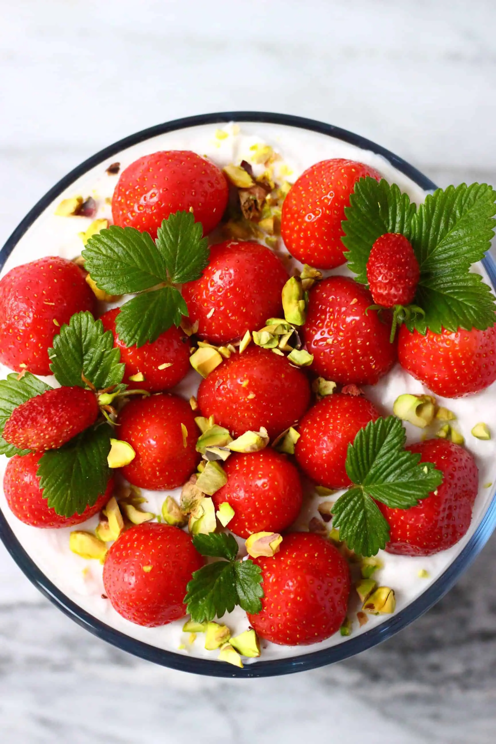 A vegan strawberry trifle in a glass trifle dish topped with fresh strawberries, green leaves and chopped pistachios