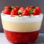 A vegan strawberry trifle with red strawberry jelly topped with yellow custard, white cream and fresh strawberries