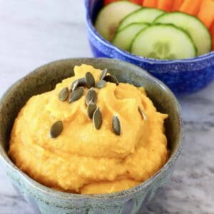 A bowl of orange pumpkin hummus topped with pumpkin seeds and a bowl of chopped vegetables