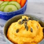 Photo of a grey bowl of orange pumpkin hummus topped with green pumpkin seeds and a blue bowl of chopped vegetables on a marble surface