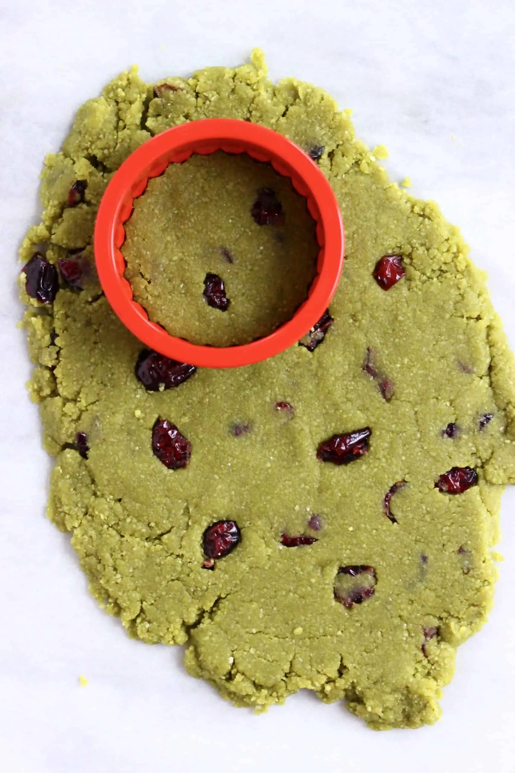 Raw vegan matcha shortbread cookie dough with dried cranberries rolled out on a baking sheet with a round cookie cutter on it