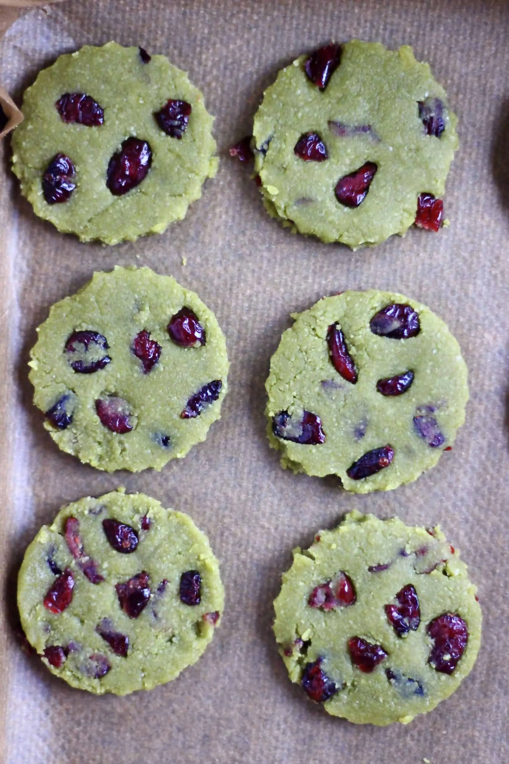 Six raw vegan matcha shortbread cookies with dried cranberries on a baking tray lined with baking paper