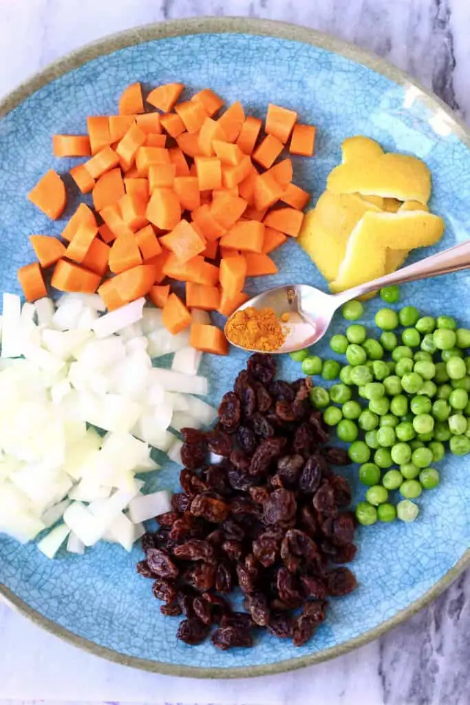 Photo of diced carrots, onions, green peas, lemon peel, raisins and turmeric on a blue plate shot from above