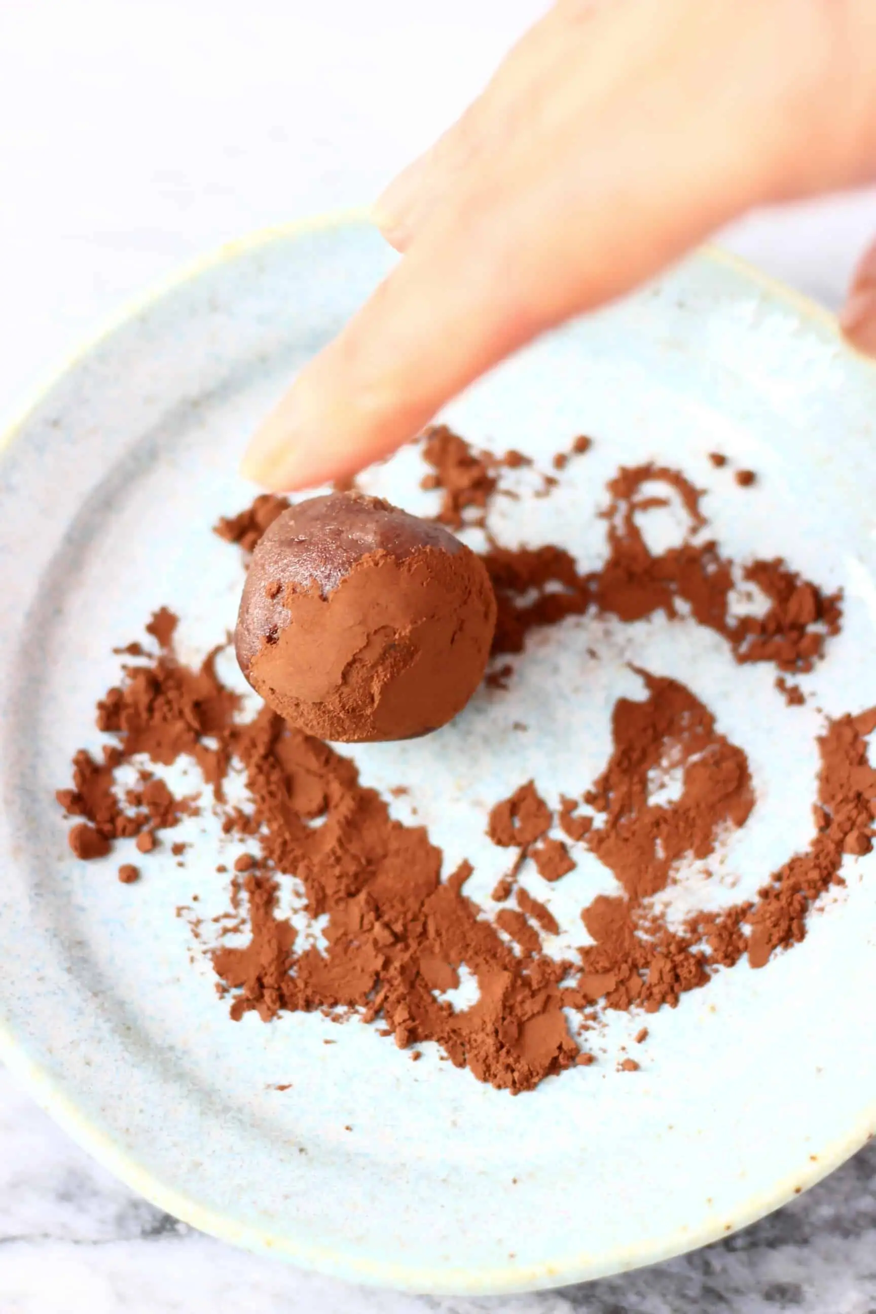 A vegan chocolate truffle being rolled in cocoa powder on a plate with fingers