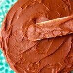 A chocolate cake on a green cake stand with a silver palette knife spreading chocolate buttercream on top of it