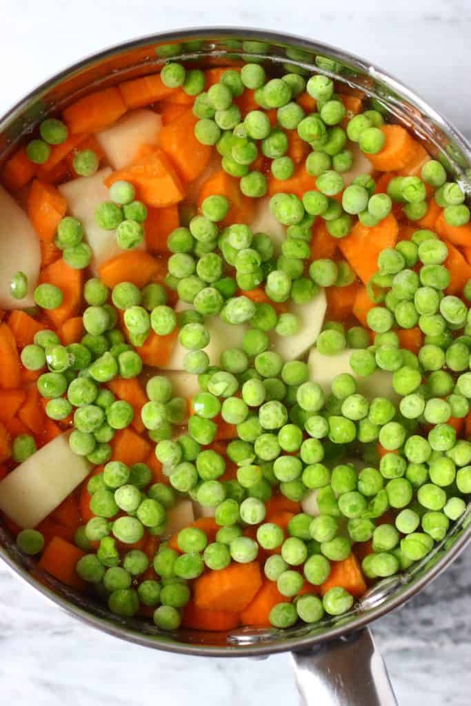 This Vegan Potato Salad is a great make-ahead side dish - it's easy to make, irresistibly creamy, and healthier than the traditional version! Made with carrots and green peas and is highly customisable. Vegetarian, dairy-free, egg-free, gluten-free, oil-free. #rhiansrecipes #vegan #potatosalad #sidedish #glutenfree #dairyfree Diced potatoes, carrots and green peas in a silver saucepan filled with water against a marble background