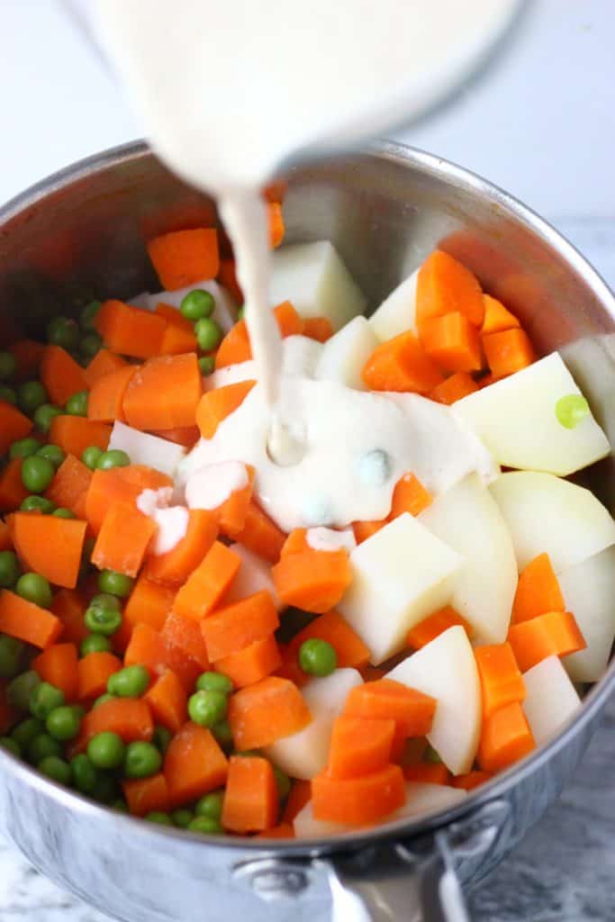 Cooked diced potatoes, carrots and green peas in a silver pan against a marble background with a jug pouring cream into it