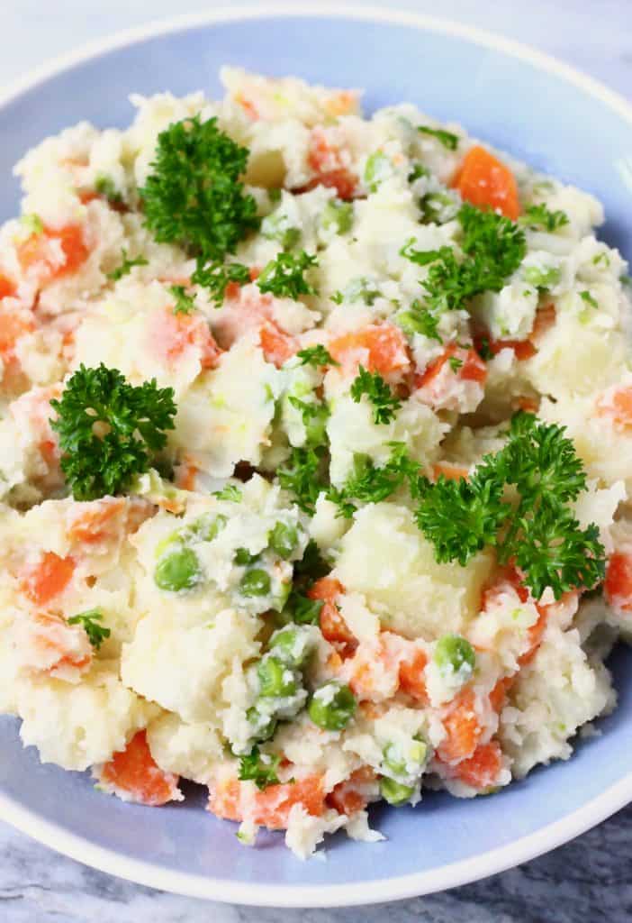 This Vegan Potato Salad is a great make-ahead side dish - it's easy to make, irresistibly creamy, and healthier than the traditional version! Made with carrots and green peas and is highly customisable. Vegetarian, dairy-free, egg-free, gluten-free, oil-free. #rhiansrecipes #vegan #potatosalad #sidedish #glutenfree #dairyfree Potato salad with carrots, green peas and mayonnaise in a blue bowl against a marble background
