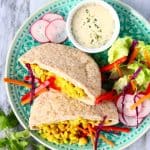 Two pittas filled with crushed chickpeas and salad on a green plate with a small white bowl of tahini sauce against a marble background