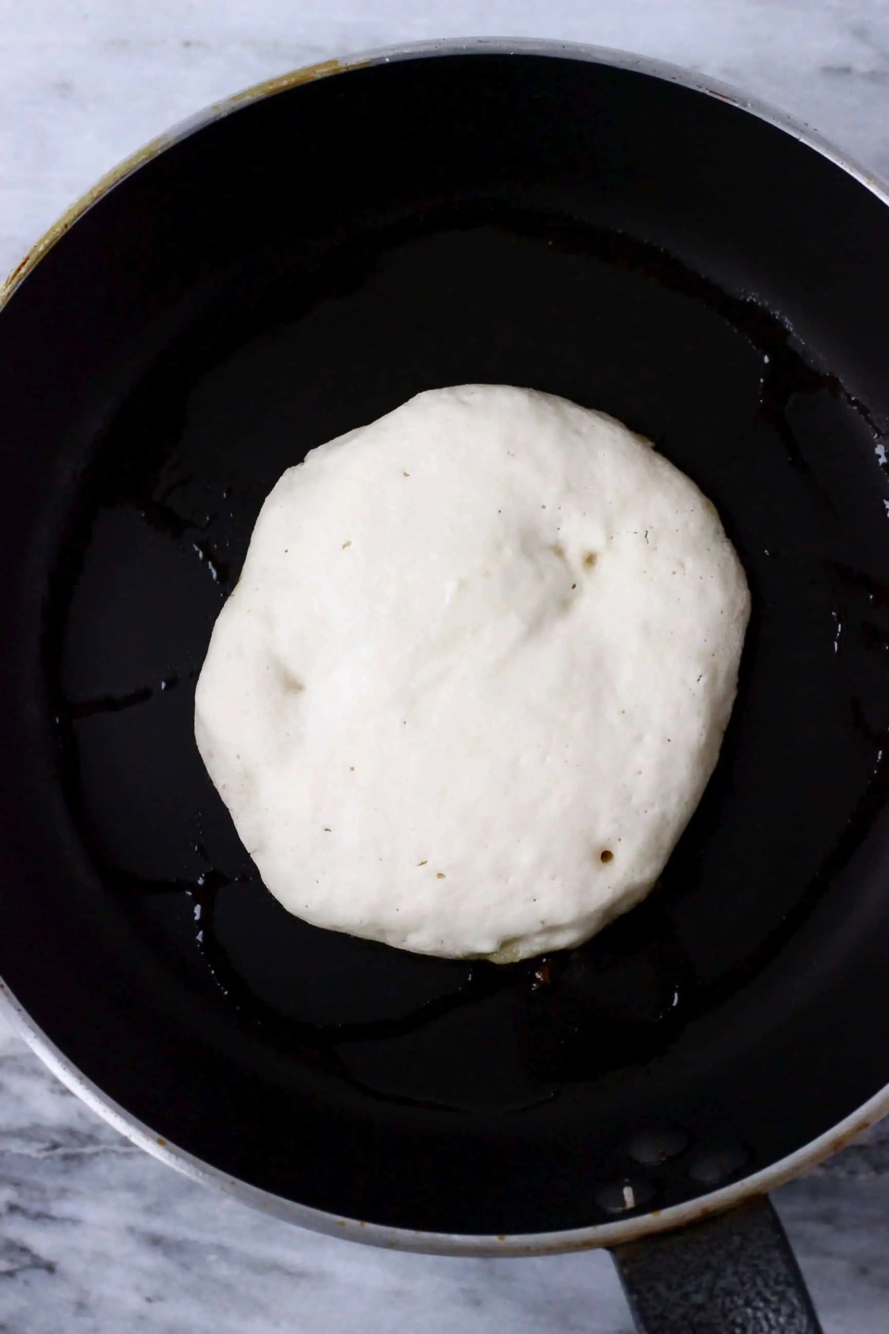 A white gluten-free vegan pancake being cooked in a black frying pan