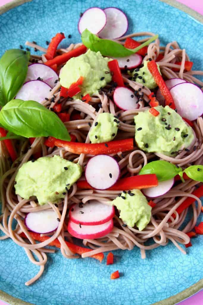 Soba noodles with edamame pesto, sliced radishes, red pepper matchsticks and basil on a blue plate against a pink background