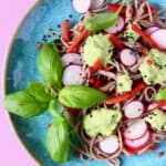 Soba noodles with edamame pesto, sliced radishes, red pepper matchsticks and basil on a blue plate against a pink background