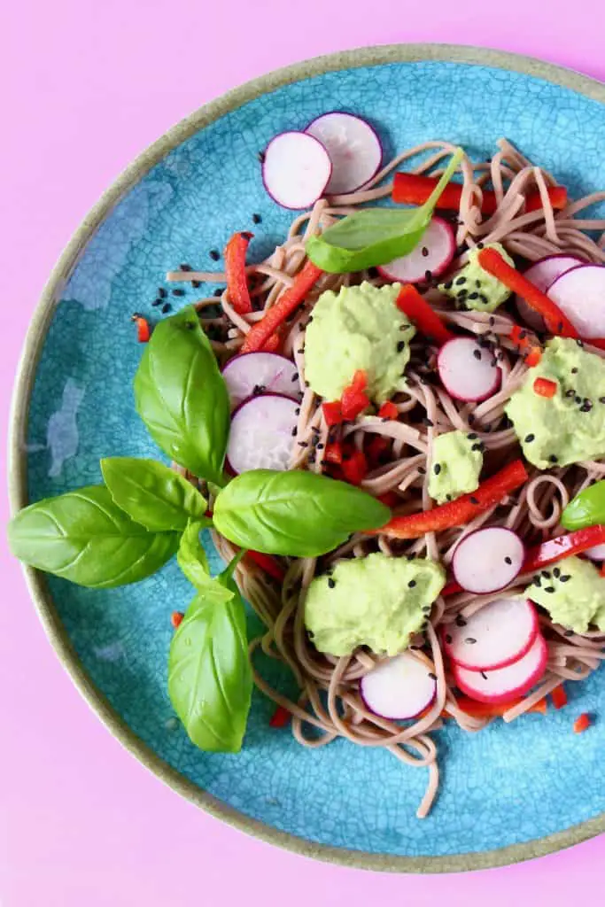 Soba noodles with edamame pesto, sliced radishes, red pepper matchsticks and basil on a blue plate against a pink background
