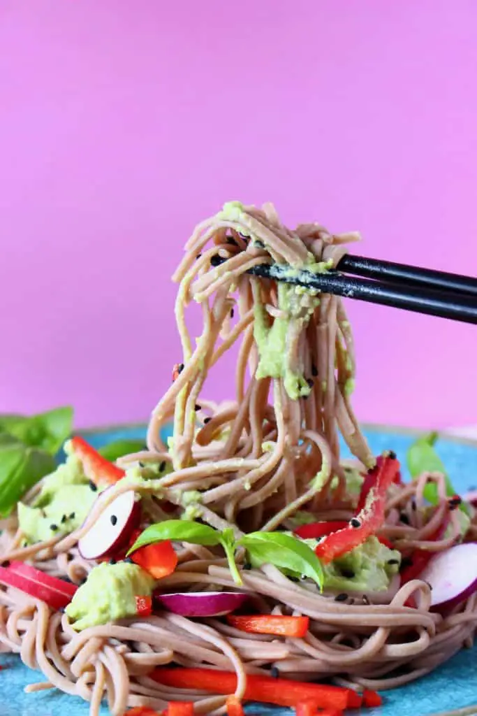 Soba noodles with edamame pesto, sliced radishes and red pepper matchsticks on a blue plate against a pink background with a pair of black chopsticks lifting up a mouthful