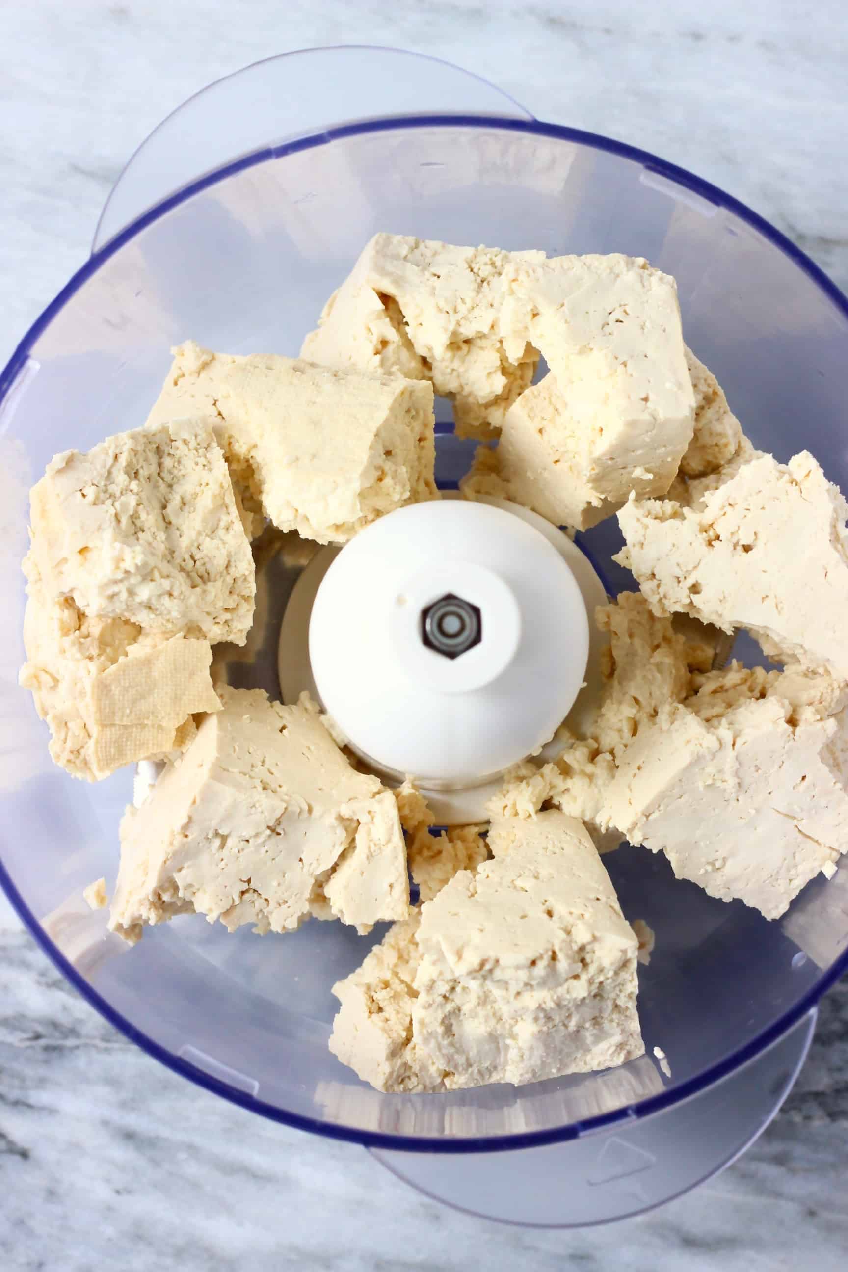 Broken up pieces of tofu in a food processor against a marble background