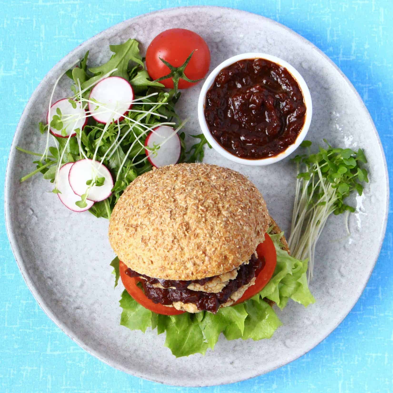 A tofu burger in a bun with brown sauce and salad on a plate