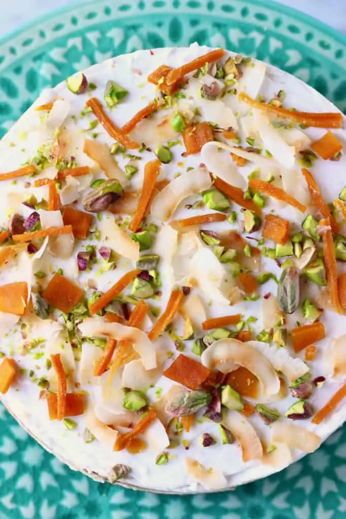 A carrot cake topped with white frosting decorated with dried mango, chopped pistachios and coconut flakes on a green cake stand