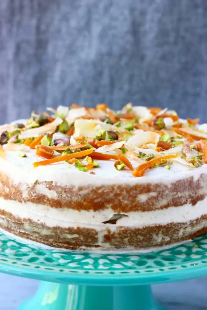 A carrot cake with white frosting decorated with dried mango, chopped pistachios and coconut flakes on a green cake stand against a grey background