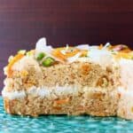 A carrot cake sponge cake with white frosting decorated with dried mango, chopped pistachios and coconut flakes on a green cake stand against a dark brown background