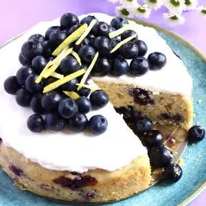 A gluten-free vegan lemon blueberry cake topped with cream and blueberries with a slice cut out of it