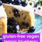 Photo of a blueberry cake topped with white frosting and fresh blueberries with a slice taken out of it and white flowers in the background