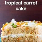 A carrot cake sponge cake with white frosting decorated with dried mango, chopped pistachios and coconut flakes on a green cake stand against a dark brown background