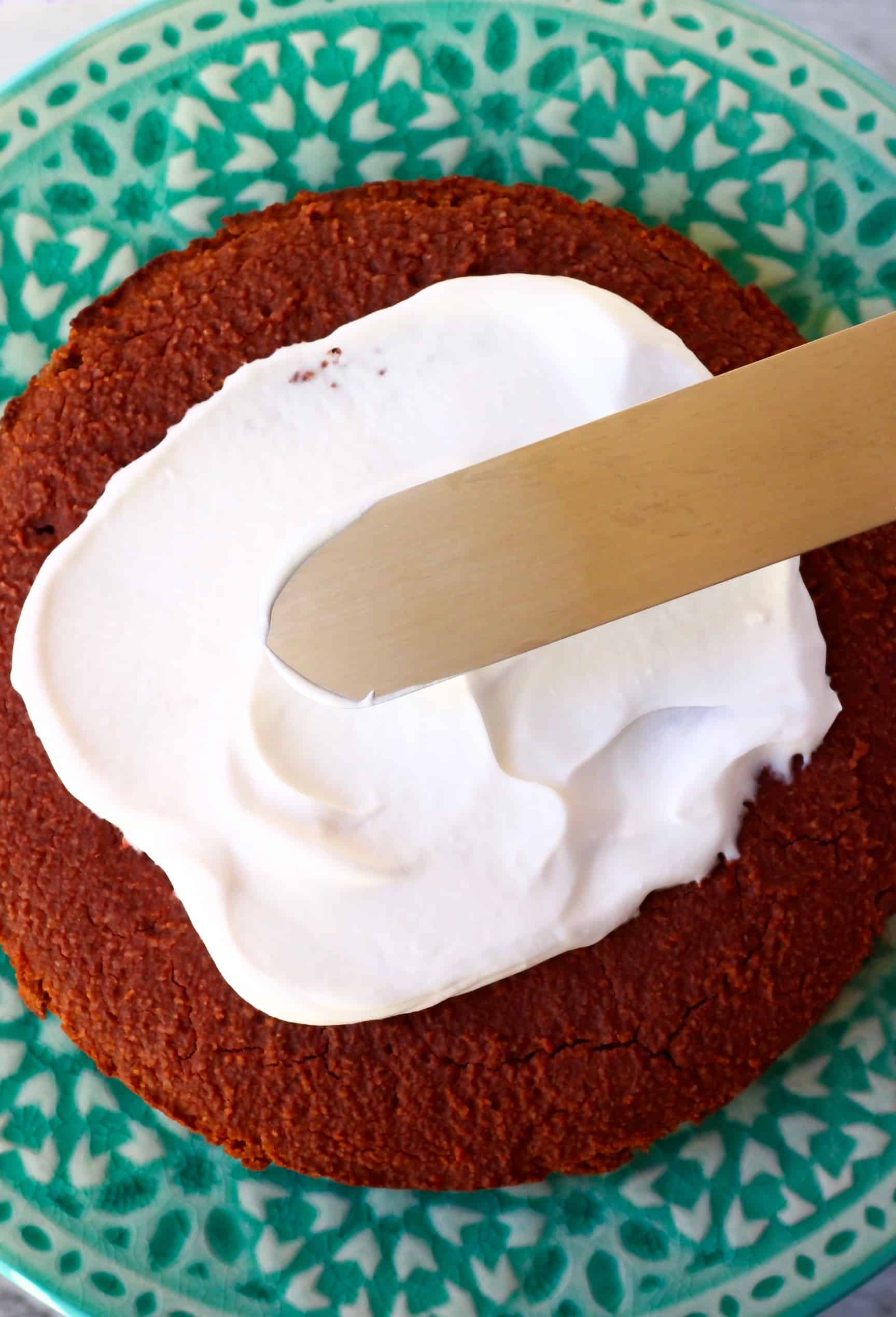 A gluten-free vegan red velvet cake sponge on a cake stand with vegan cream cheese frosting being spread on it with a palette knife