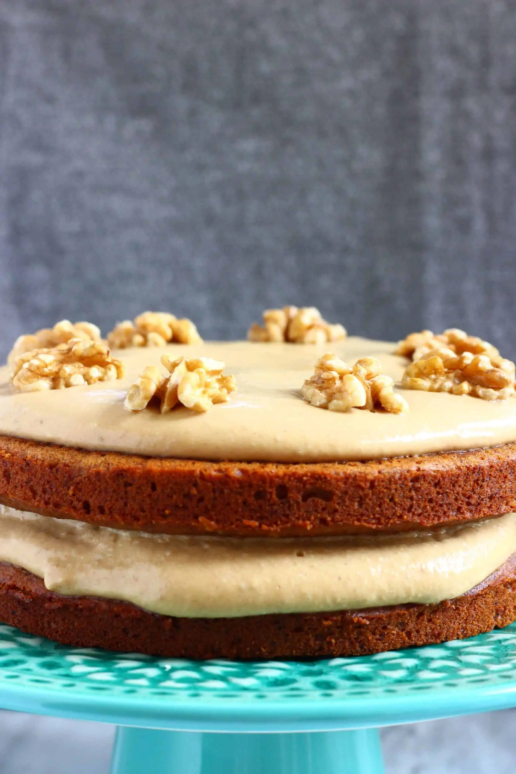 A gluten-free vegan coffee cake sandwiched with coffee frosting topped with walnuts on a cake stand