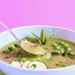 Green soup topped with green vegetables in a light blue bowl against a pink background with a silver spoon lifting up a mouthful