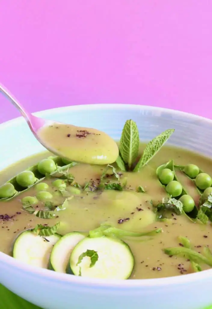 Green soup topped with green vegetables in a light blue bowl against a pink background with a silver spoon lifting up a mouthful