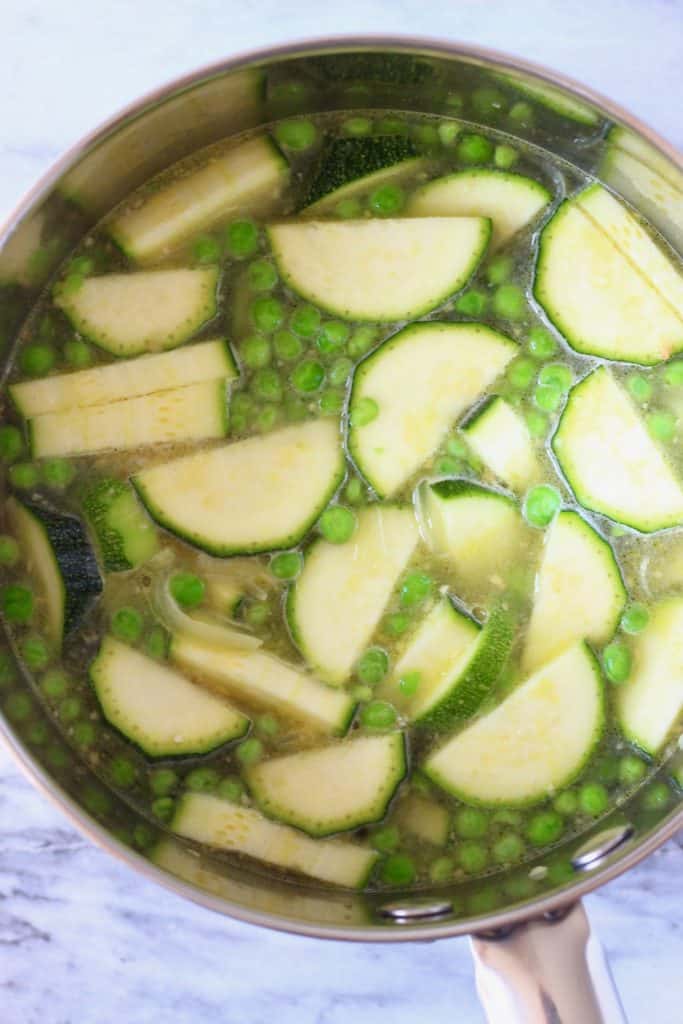 Sliced courgettes, peas and water in a silver pan against a marble background