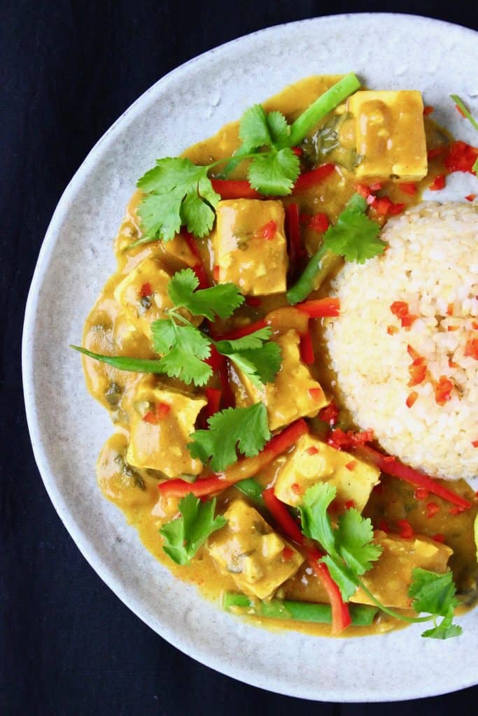 Cubes of tofu in a golden brown curry sauce with French beans and sliced red pepper with a pile of rice on a grey plate against a black background