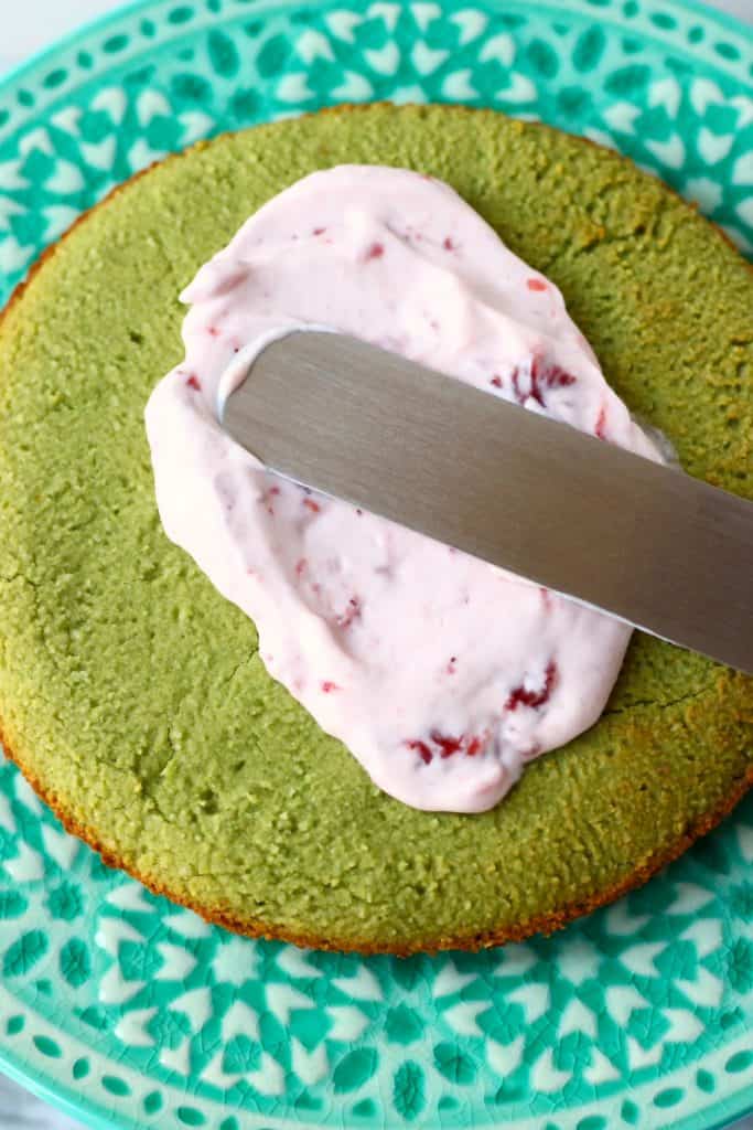 A matcha sponge cake with strawberry frosting being spread on it using a palette knife