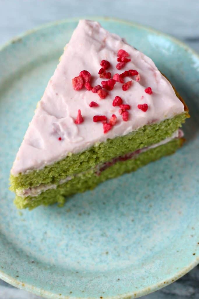 A slice of matcha sponge cake sandwiched with strawberry frosting on a blue plate against a marble background